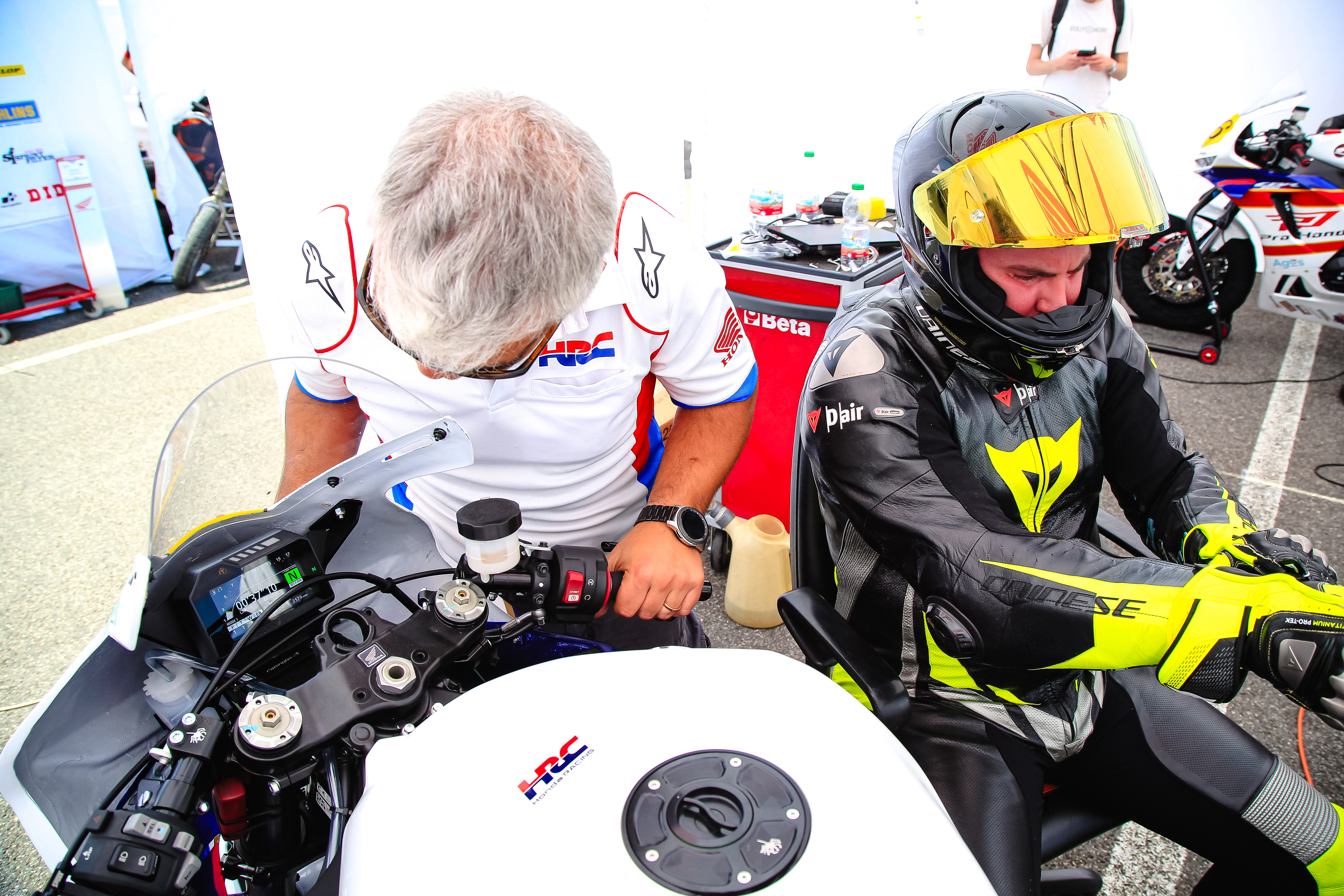 Two motorcycle racers preparing for a race. One rider in a white shirt leans over a Honda racing bike, while another in a black and yellow suit sits on a different motorcycle. Racing equipment and other bikes are visible in the background.